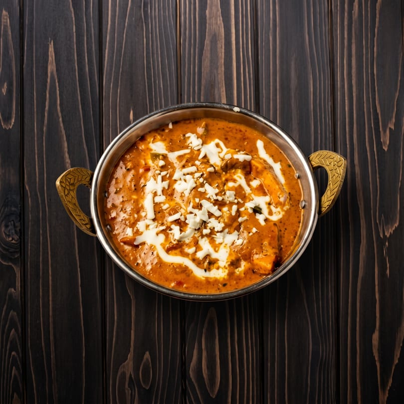 Rich orange paneer curry with cream and cheese in a decorative metal bowl on dark wood.