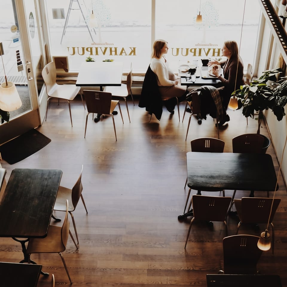Cafe interior with two women dining at a window table, wooden floors, and warm lighting.