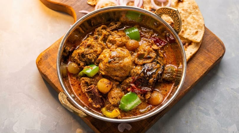 A metal bowl of chicken curry with onions and bell peppers, served with flatbread on a wooden board.