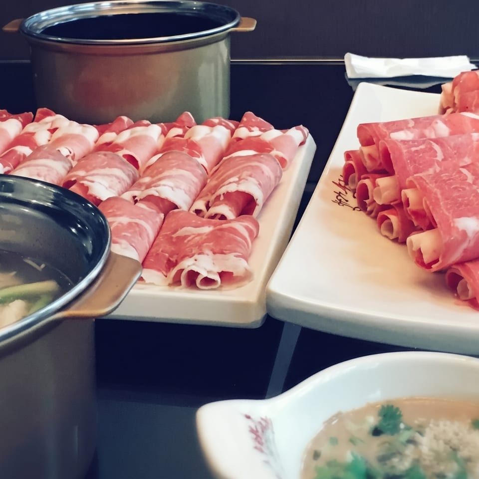 Hot pot meal with two pots, plates of thinly sliced raw meat, and a dipping sauce.