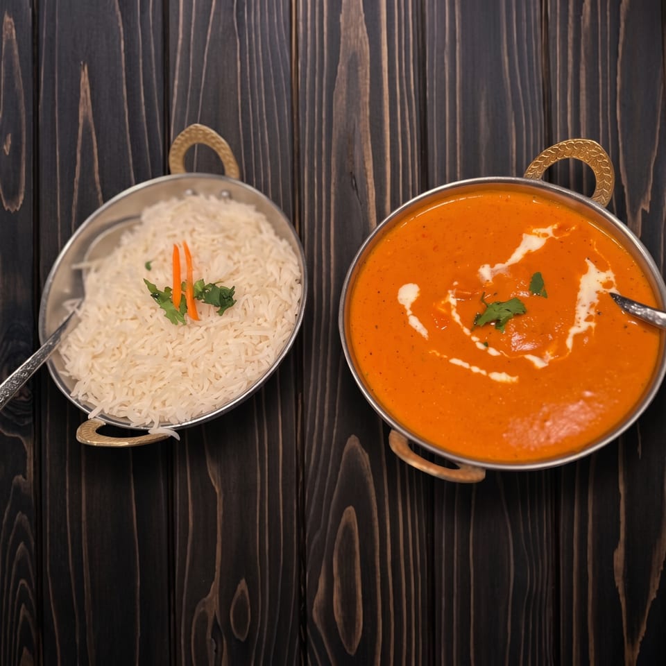 Orange curry with cream swirl and white rice, served in metal bowls on a dark wooden surface.