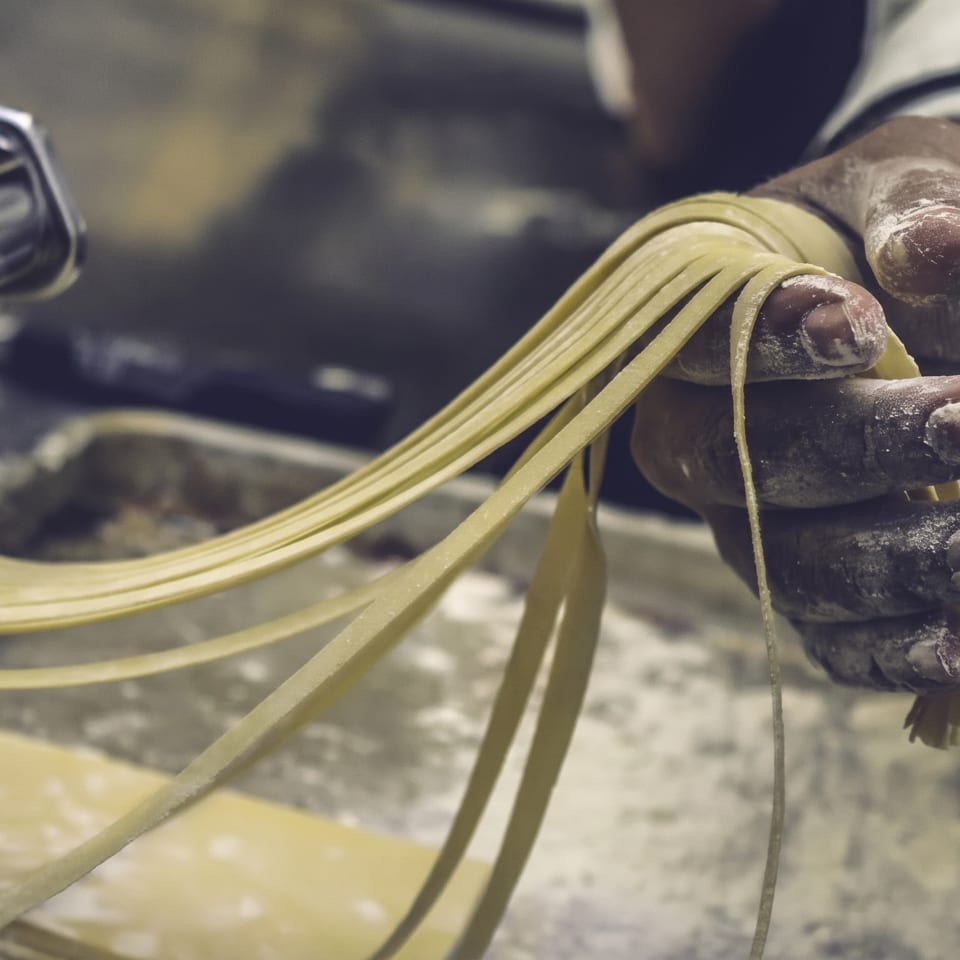 Chef's flour-dusted hands pull fresh pasta strands from a pasta machine.