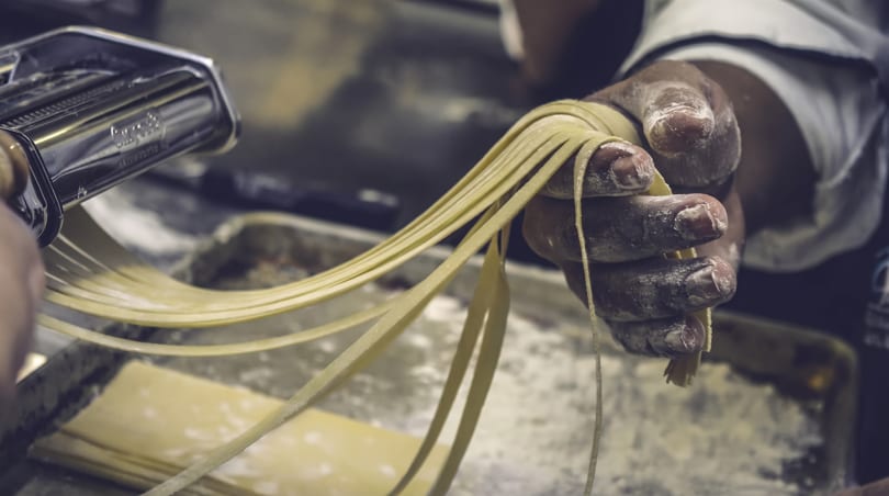 Chef's flour-dusted hands pull fresh pasta strands from a pasta machine.