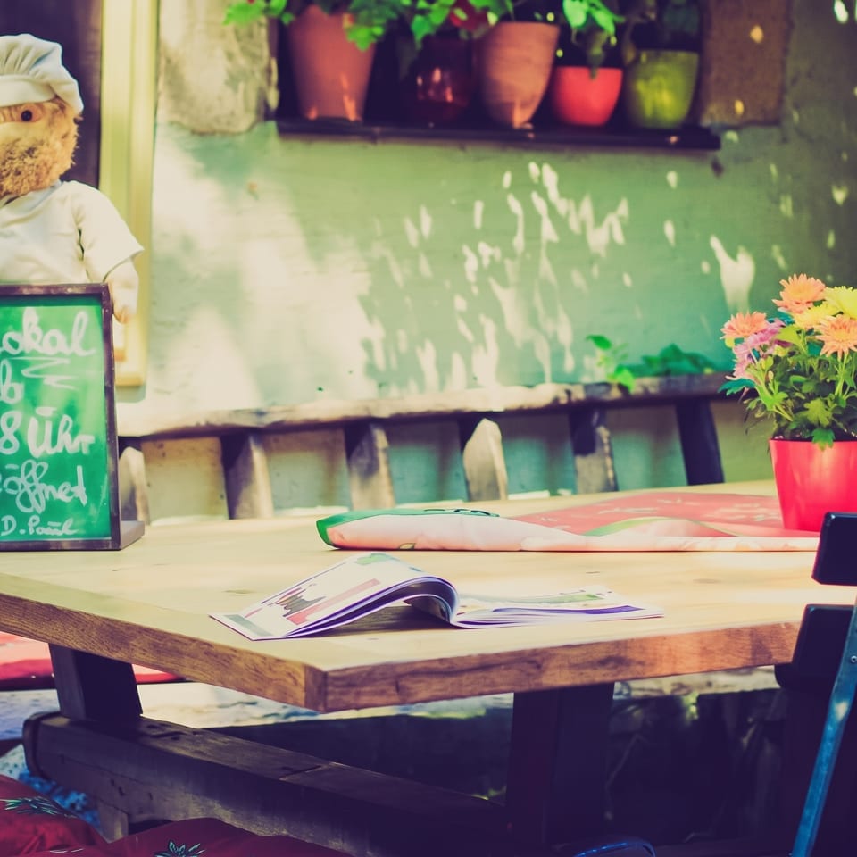 Sunny outdoor restaurant patio with a wooden table, chairs, flowers, and a chef doll by a chalkboard.